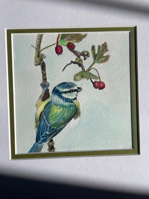 A framed watercolor painting of a bluebird perched on a branch with red berries, displayed against a light gray wall with soft shadows.